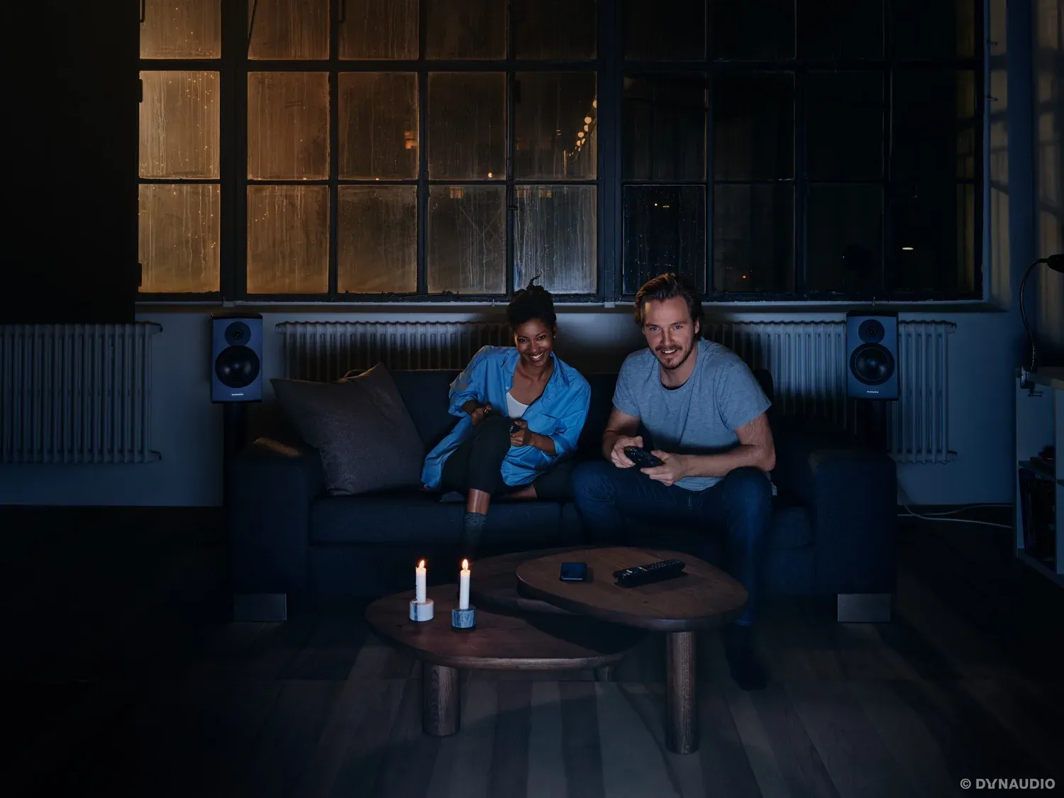 Paar spielt auf dem Sofa Videospiele, im Hintergrund Regallautsprecher