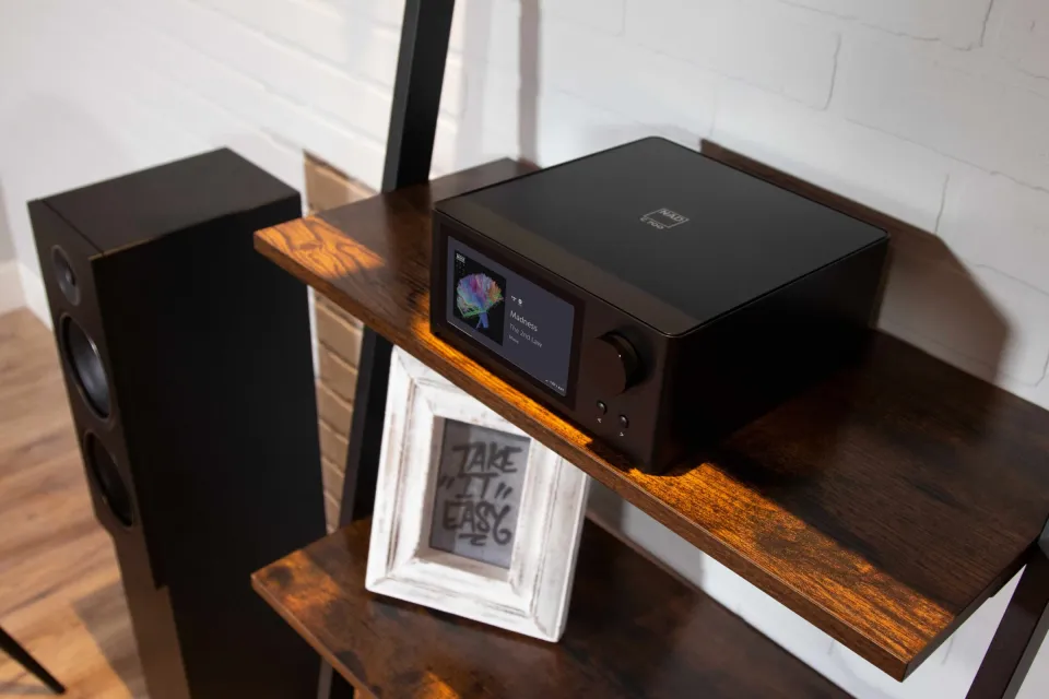 NAD-Gerät mit Farbdisplay auf einem Holzregal neben einem Lautsprecher